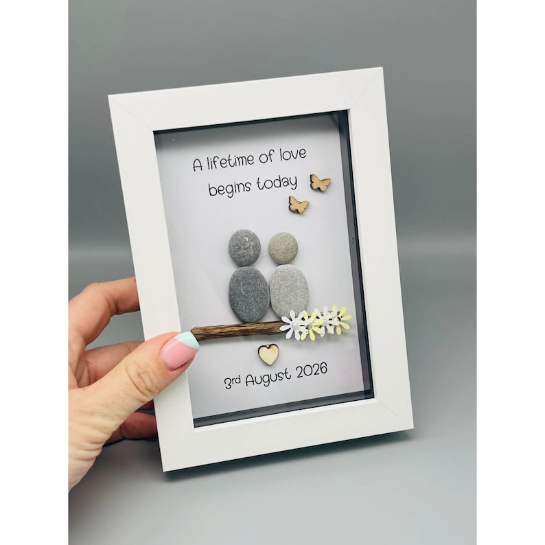 May include: A framed pebble art piece with the text "A lifetime of love begins today" and the date "3rd August 2026". The artwork depicts a couple made of pebbles, with wooden butterflies and flowers, in a white frame.