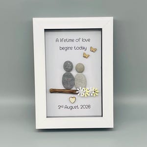 May include: Framed artwork with the text "A lifetime of love begins today." The image shows two pebble figures, wooden butterflies, and a heart. The date "3rd August 2026" is included. The artwork is a unique, handcrafted piece, presented in a white frame.