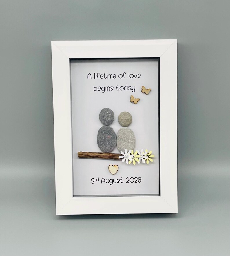 May include: Framed artwork with the text "A lifetime of love begins today." The image shows two pebble figures, wooden butterflies, and a heart. The date "3rd August 2026" is included. The artwork is a unique, handcrafted piece, presented in a white frame.