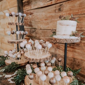 May include: A rustic wedding cake display with a white cake on a wooden stand and a tiered cake stand filled with cake pops. The cake pops are decorated with white, brown, and light blue frosting. The cake pops are on wooden sticks and are arranged in a circle on the cake stand. The cake stand is made of wood and has a rustic look. The cake pops are surrounded by greenery.