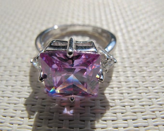 Puede incluir: Anillo plateado con una gran gema de color púrpura claro de corte cuadrado. Dos gemas claras más pequeñas flanquean la piedra principal. El anillo está sobre una superficie texturizada blanquecina.