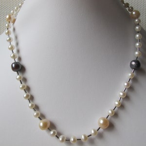 May include: A pearl necklace with a mix of white, cream, and dark gray pearls. The necklace is strung on a thin, silver-colored wire and is displayed on a white bust.