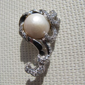 May include: A silver-toned brooch featuring a large, round, cream-colored pearl at its center. The pearl is surrounded by a swirling, ornate design embellished with small, clear crystals. The brooch has a pin closure on the back.