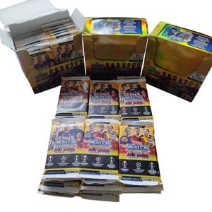 Puede incluir: Múltiples paquetes de cromos Match Attax, con jugadores de fútbol. Los paquetes están en cajas y muestran el texto "STAGIONE 2021/22 12 CARD €2". Las cartas son para la Champions League y la Europa League.