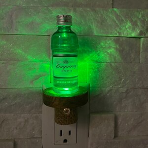 May include: A green glass bottle of Tanqueray London Dry Gin with a cork stopper, mounted on a wooden base and plugged into a white electrical outlet. The bottle is illuminated from within, casting a green glow.