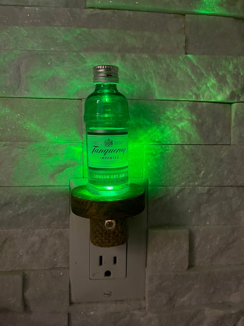May include: A green glass bottle of Tanqueray London Dry Gin with a cork stopper, mounted on a wooden base and plugged into a white electrical outlet. The bottle is illuminated from within, casting a green glow.