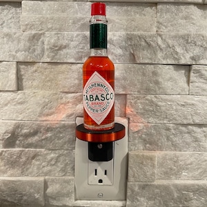 May include: A red glass bottle of Tabasco brand pepper sauce is mounted on a white electrical outlet with a black faceplate. The bottle is labeled "McIlhenny Co. Avery Island, LA. Tabasco Brand Pepper Sauce 2 fl. oz. (59 ml)."