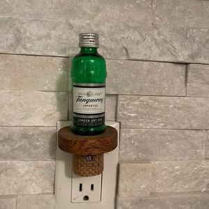 May include: A small bottle of Tanqueray London Dry Gin, 50 ml, is displayed on a wooden bottle stopper, which is plugged into a standard electrical outlet. The bottle is green and has a silver cap. The outlet is white.