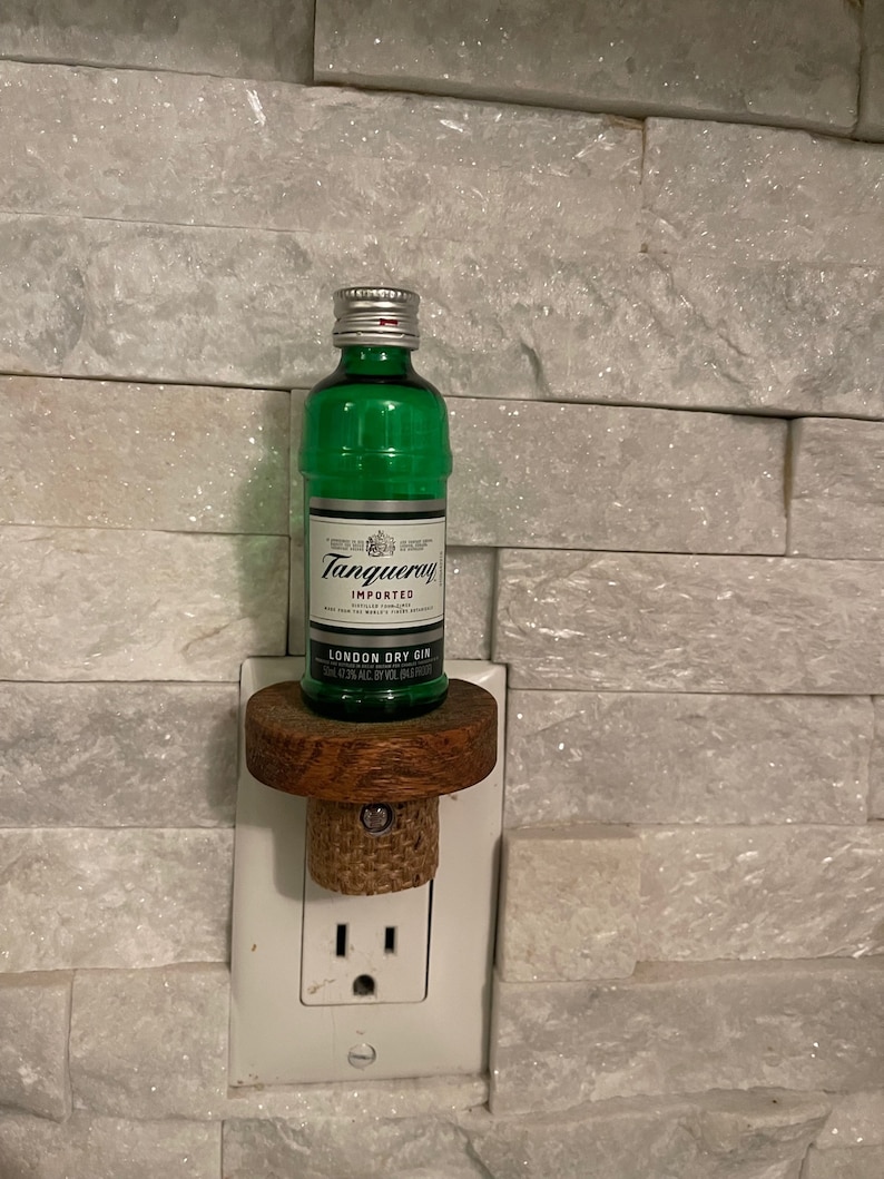 May include: A small bottle of Tanqueray London Dry Gin, 50 ml, is displayed on a wooden bottle stopper, which is plugged into a standard electrical outlet. The bottle is green and has a silver cap. The outlet is white.