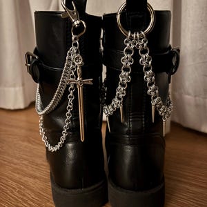 May include: Pair of black ankle boots with silver chain and cross charms. The boots have buckles and a chunky sole. One boot has a chain with a cross pendant, the other has a chain with spikes.