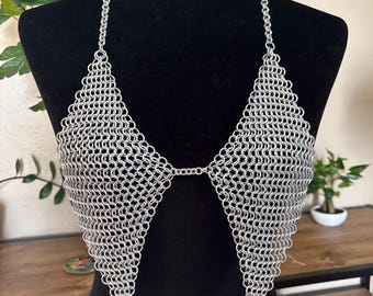 May include: A silver chainmail top with a halter neck design. The top is made of small, interlocking metal rings, creating a mesh-like appearance. The design features triangular cups and decorative metal spikes at the bottom.