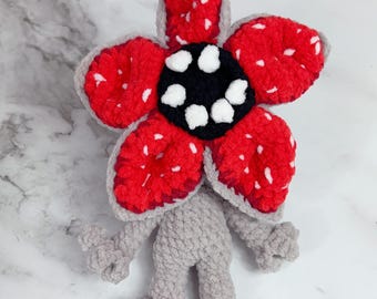 May include: A crochet plush toy featuring a gray humanoid body with thin arms and legs, topped with a large, five-petaled flower head. The petals are bright red with white speckles, surrounding a black center with white circular teeth.