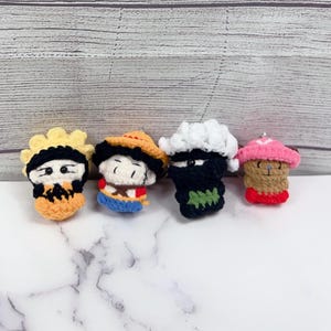 May include: Four small, handmade crochet figures in a row. The figures are in the shape of cartoon characters, each with unique colors and designs. The first is yellow and orange, the second has a straw hat, the third is black and white, and the fourth is pink and brown.