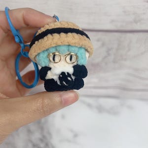 K&ouml;nnte beinhalten: Handgefertigter H&auml;kel-Schl&uuml;sselanh&auml;nger mit einer kleinen Figur mit hellblauer Frisur, goldgerahmter Brille und einem beigefarbenen Hut mit schwarzen Streifen. Die Figur tr&auml;gt ein schwarzes Outfit. Ein blauer Schl&uuml;sselanh&auml;ngerring ist befestigt.