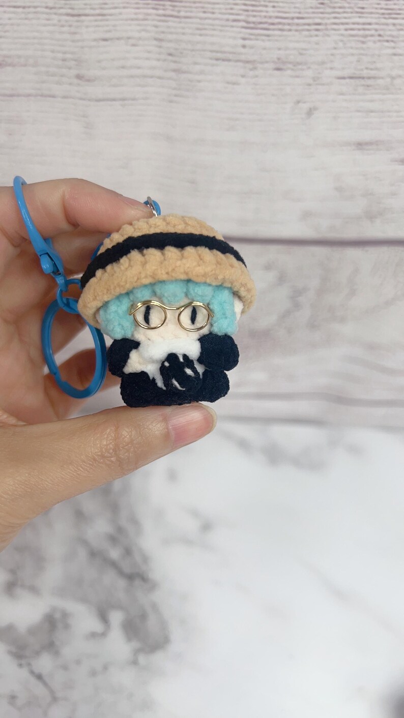 K&ouml;nnte beinhalten: Handgefertigter H&auml;kel-Schl&uuml;sselanh&auml;nger mit einer kleinen Figur mit hellblauer Frisur, goldgerahmter Brille und einem beigefarbenen Hut mit schwarzen Streifen. Die Figur tr&auml;gt ein schwarzes Outfit. Ein blauer Schl&uuml;sselanh&auml;ngerring ist befestigt.