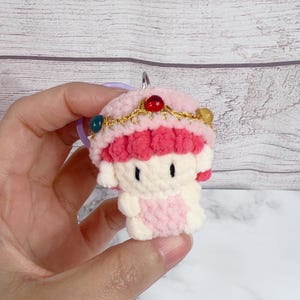 K&ouml;nnte beinhalten: Ein handgefertigter H&auml;kel-Schl&uuml;sselanh&auml;nger mit einer kleinen, puppenartigen Figur in Rosa- und Wei&szlig;t&ouml;nen. Die Figur hat einen rosa Hut mit dekorativen Perlen und einer goldfarbenen Krone. Der Schl&uuml;sselanh&auml;nger wird von einem silbernen Ring gehalten.