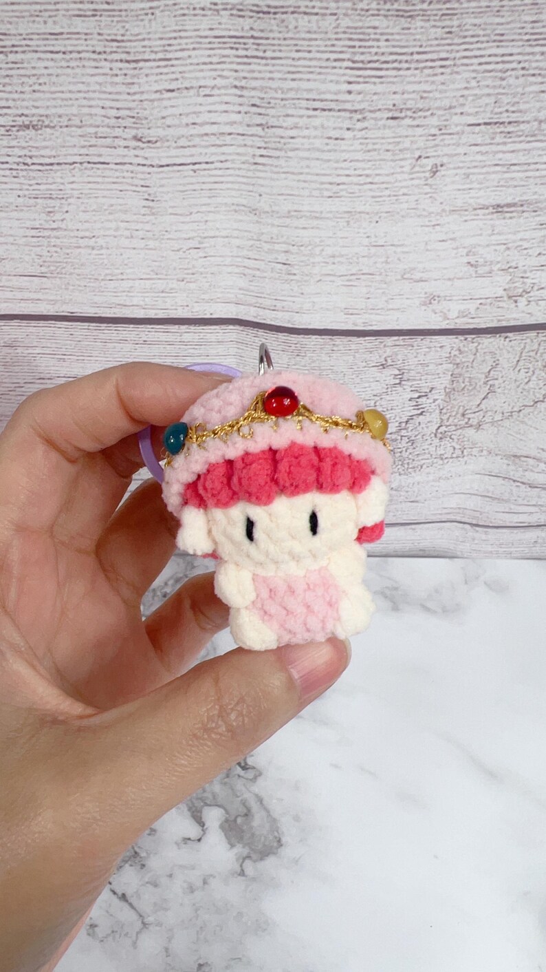 K&ouml;nnte beinhalten: Ein handgefertigter H&auml;kel-Schl&uuml;sselanh&auml;nger mit einer kleinen, puppenartigen Figur in Rosa- und Wei&szlig;t&ouml;nen. Die Figur hat einen rosa Hut mit dekorativen Perlen und einer goldfarbenen Krone. Der Schl&uuml;sselanh&auml;nger wird von einem silbernen Ring gehalten.