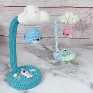 May include: Two crochet lamps with cloud-shaped tops and whale or sea creature pendants. One lamp is turquoise with a blue whale, the other is pastel with a pink whale. The bases are decorated with sea-themed embellishments.