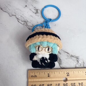 K&ouml;nnte beinhalten: Handgefertigter H&auml;kel-Schl&uuml;sselanh&auml;nger mit einer kleinen Figur mit blauen Haaren, Brille und einem schwarz-beigen Hut. Die Figur tr&auml;gt ein schwarzes Outfit mit wei&szlig;em Kragen. Der Schl&uuml;sselanh&auml;nger ist etwa 2,5 cm gro&szlig;.