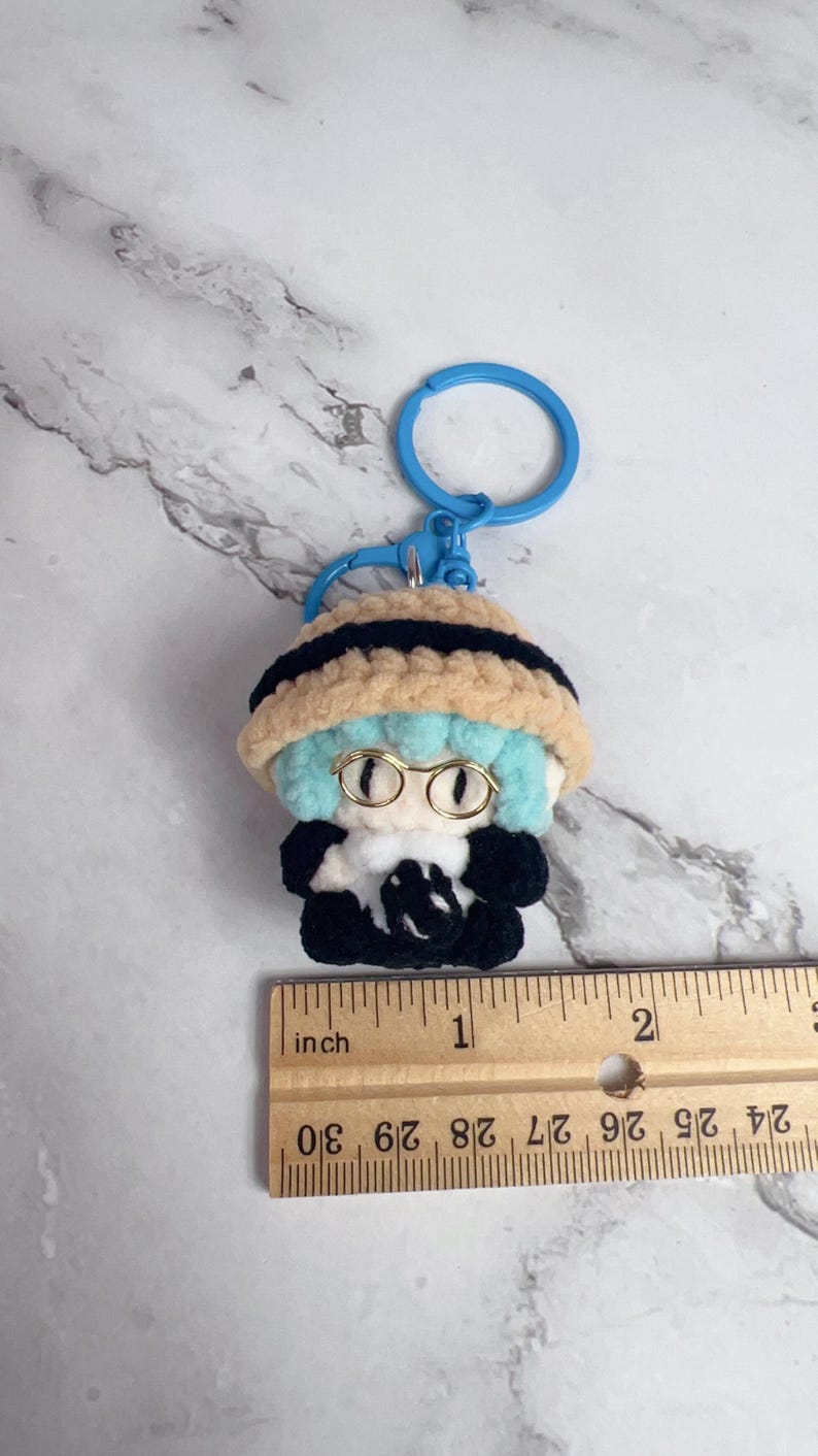 K&ouml;nnte beinhalten: Handgefertigter H&auml;kel-Schl&uuml;sselanh&auml;nger mit einer kleinen Figur mit blauen Haaren, Brille und einem schwarz-beigen Hut. Die Figur tr&auml;gt ein schwarzes Outfit mit wei&szlig;em Kragen. Der Schl&uuml;sselanh&auml;nger ist etwa 2,5 cm gro&szlig;.