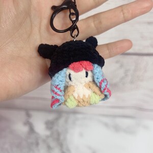 K&ouml;nnte beinhalten: Ein handgefertigter H&auml;kel-Schl&uuml;sselanh&auml;nger mit einer kleinen, skurrilen Figur mit schwarzem Hut, blau-rosa Haaren und einem cremefarbenen K&ouml;rper. Der Schl&uuml;sselanh&auml;nger hat einen schwarzen Verschluss und Ring.