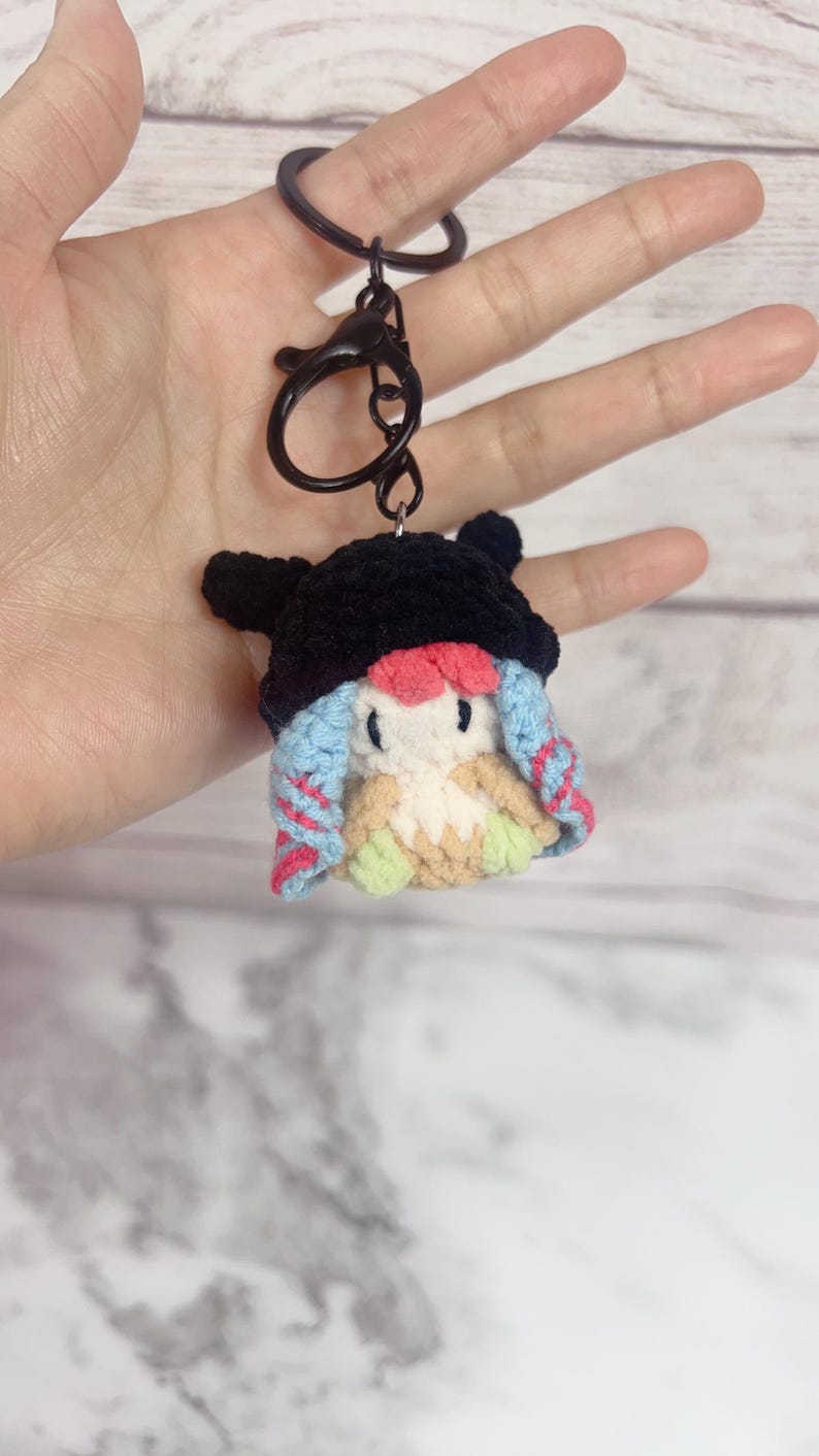 K&ouml;nnte beinhalten: Ein handgefertigter H&auml;kel-Schl&uuml;sselanh&auml;nger mit einer kleinen, skurrilen Figur mit schwarzem Hut, blau-rosa Haaren und einem cremefarbenen K&ouml;rper. Der Schl&uuml;sselanh&auml;nger hat einen schwarzen Verschluss und Ring.