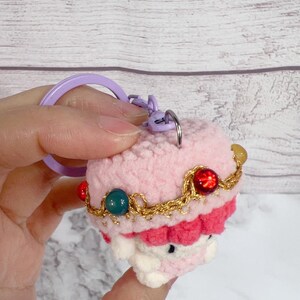 K&ouml;nnte beinhalten: Ein handgefertigter H&auml;kel-Schl&uuml;sselanh&auml;nger in Form eines rosa Cupcakes, verziert mit einer goldfarbenen Kette und bunten Perlen. Der Schl&uuml;sselanh&auml;nger hat einen lila Ring und Verschluss. Das Cupcake-Design hat eine wei&szlig;e Basis und eine rote Frosting-Schicht.