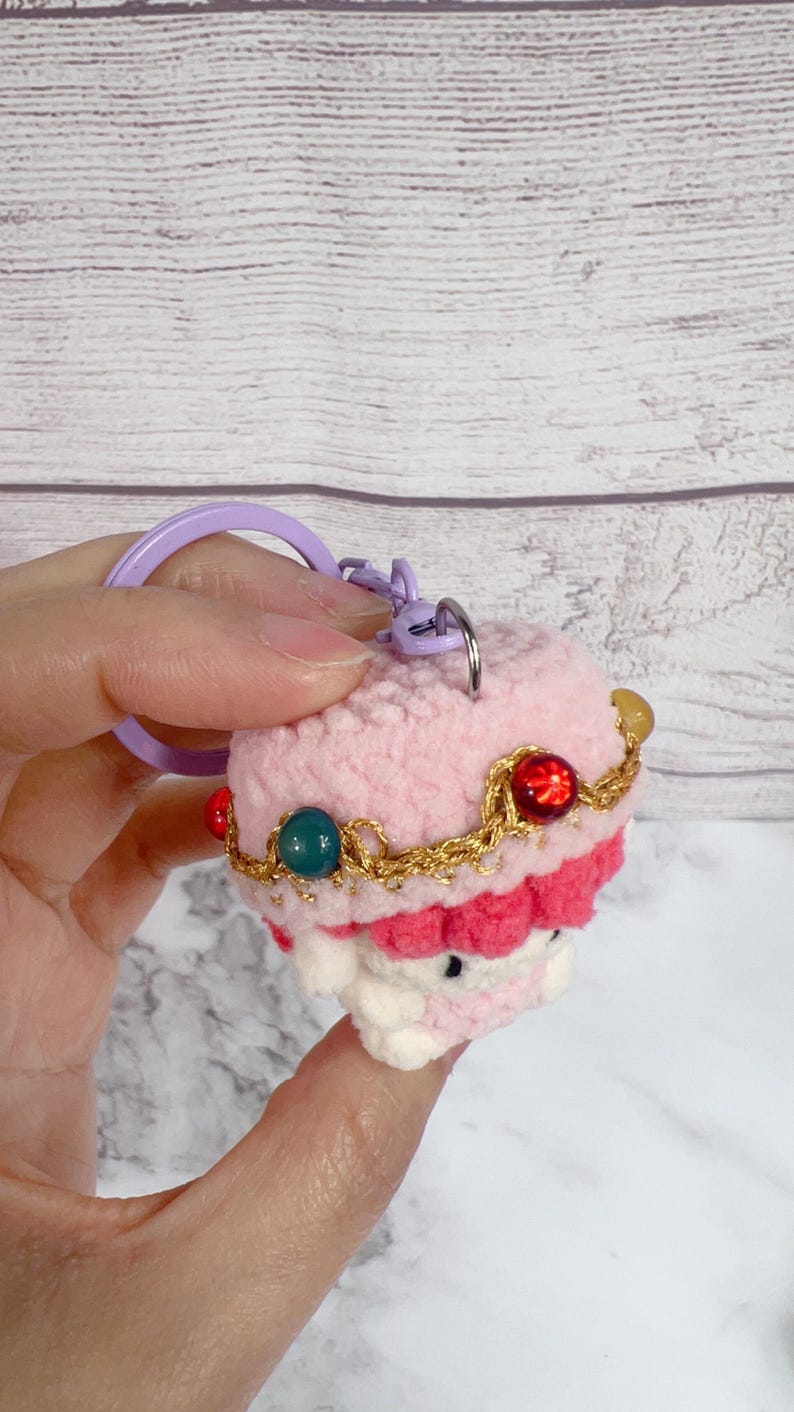 K&ouml;nnte beinhalten: Ein handgefertigter H&auml;kel-Schl&uuml;sselanh&auml;nger in Form eines rosa Cupcakes, verziert mit einer goldfarbenen Kette und bunten Perlen. Der Schl&uuml;sselanh&auml;nger hat einen lila Ring und Verschluss. Das Cupcake-Design hat eine wei&szlig;e Basis und eine rote Frosting-Schicht.