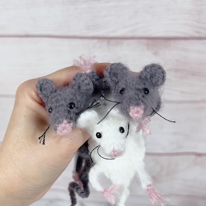 May include: Three handmade crochet mice in gray and white, held in a hand. The mice have pink noses and ears, black eyes, and long tails. The background is a light wood grain.