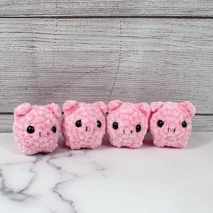 May include: Four pink crocheted pig plushies with black eyes and snouts. The plushies are lined up in a row on a white marble surface.
