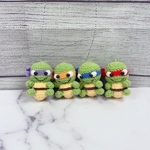May include: Four handmade crochet turtle figurines, each with a different colored mask: purple, orange, blue, and red. The turtles have green shells, tan bellies, and white eyes. They are displayed on a white marble surface against a light wood-grain background.