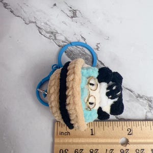 K&ouml;nnte beinhalten: Geh&auml;kelter Schl&uuml;sselanh&auml;nger mit einem skurrilen Design in Hellblau, Beige, Schwarz und Wei&szlig;. Der Schl&uuml;sselanh&auml;nger hat einen blauen Ring und einen goldfarbenen Drahtrahmen. Der Artikel ist etwa 2,5 cm lang.