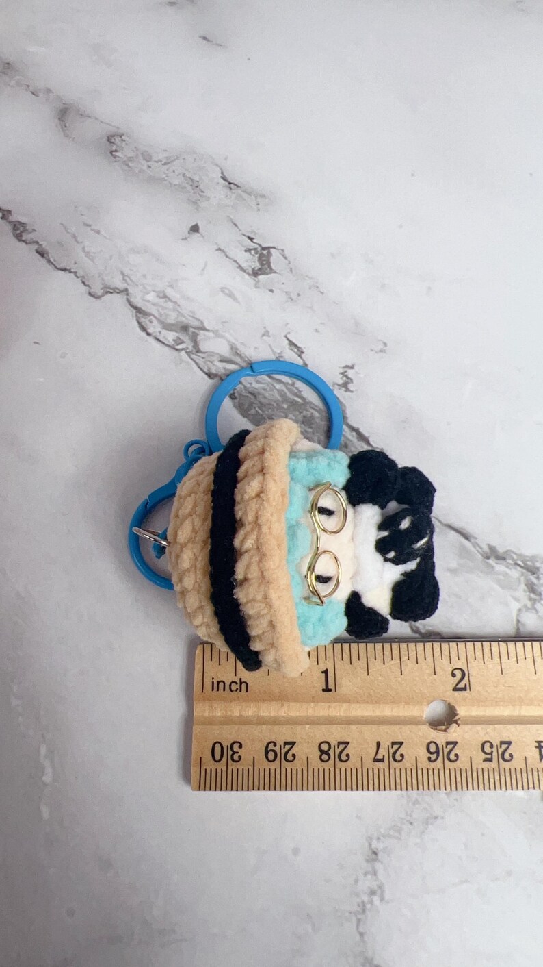 K&ouml;nnte beinhalten: Geh&auml;kelter Schl&uuml;sselanh&auml;nger mit einem skurrilen Design in Hellblau, Beige, Schwarz und Wei&szlig;. Der Schl&uuml;sselanh&auml;nger hat einen blauen Ring und einen goldfarbenen Drahtrahmen. Der Artikel ist etwa 2,5 cm lang.