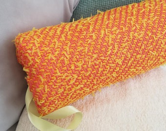 May include: A yellow and pink cylindrical cushion with a yellow ribbon tied around it. The cushion is made of a textured fabric and is sitting on a white fluffy surface.