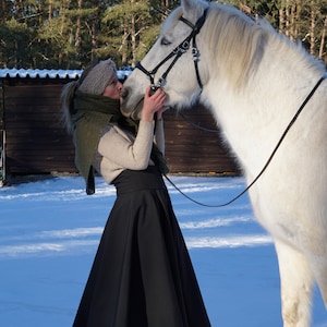 May include: A woman in a black skirt and a green scarf is kissing a white horse on the nose. The horse is wearing a black bridle and is standing in the snow.