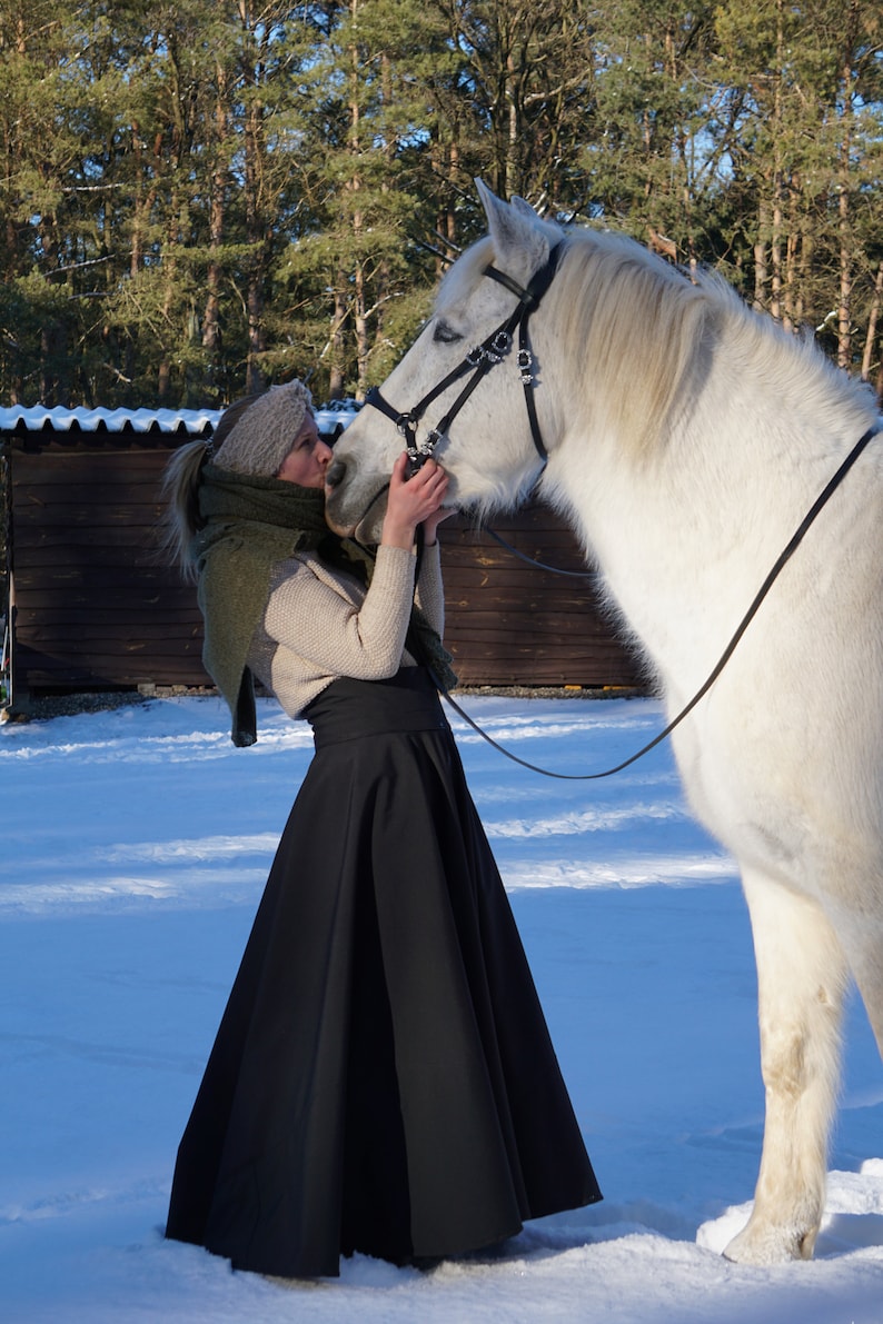 May include: A woman in a black skirt and a green scarf is kissing a white horse on the nose. The horse is wearing a black bridle and is standing in the snow.