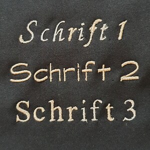 May include: Close-up of embroidered text samples on a dark background. The text reads "Schrift 1", "Schrift 2", and "Schrift 3" in a decorative font. The thread colors are light beige and off-white.