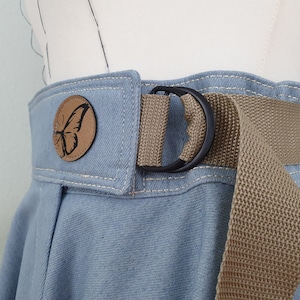 May include: Close-up of a light blue denim skirt with a tan canvas belt and a black metal D-ring buckle. A round, brown leather patch with a black butterfly design is sewn onto the skirt.