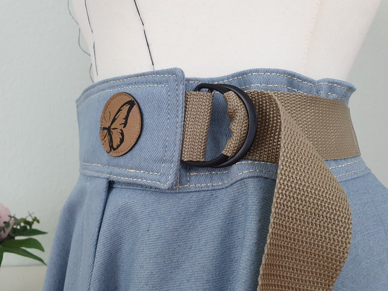 May include: Close-up of a light blue denim skirt with a tan canvas belt and a black metal D-ring buckle. A round, brown leather patch with a black butterfly design is sewn onto the skirt.