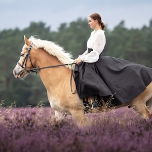 May include: A light brown horse with a white mane and tail is ridden through a field of purple flowers. The rider wears a white blouse and a long black skirt. The sky is overcast.