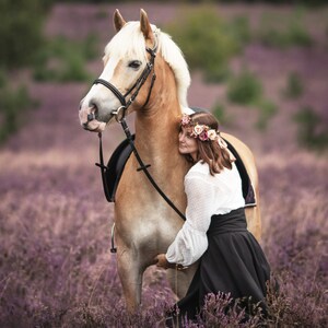 May include: A light brown horse with a white mane and tail stands in a field of purple flowers. The horse wears a bridle and saddle. A person wearing a white blouse, black skirt, and floral crown embraces the horse.