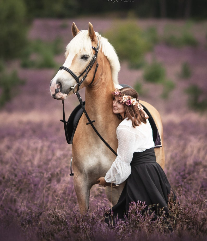 May include: A light brown horse with a white mane and tail stands in a field of purple flowers. The horse wears a bridle and saddle. A person wearing a white blouse, black skirt, and floral crown embraces the horse.