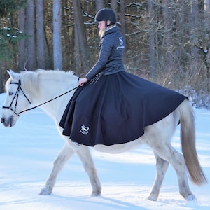 May include: A woman wearing a black riding skirt and a black jacket rides a white horse in a snowy forest. The horse is wearing a black bridle and a black saddle.