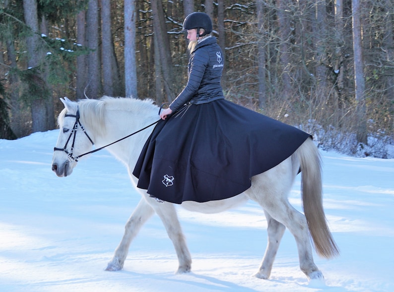 May include: A woman wearing a black riding skirt and a black jacket rides a white horse in a snowy forest. The horse is wearing a black bridle and a black saddle.
