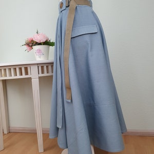 May include: A light blue skirt with a wide, flowing design. It features a matching belt with a wooden buckle and a pocket detail. The skirt is displayed on a mannequin, showcasing its shape and style.