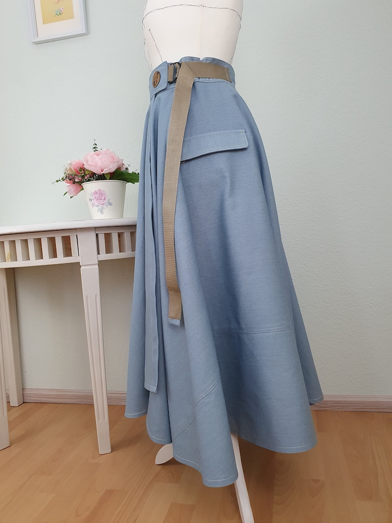 May include: A light blue skirt with a wide, flowing design. It features a matching belt with a wooden buckle and a pocket detail. The skirt is displayed on a mannequin, showcasing its shape and style.