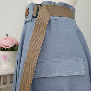 May include: Light blue denim skirt with a tan belt and a pocket detail. The skirt has a high waist design with a decorative wooden button. The tan belt is made of a woven material and has a black buckle.