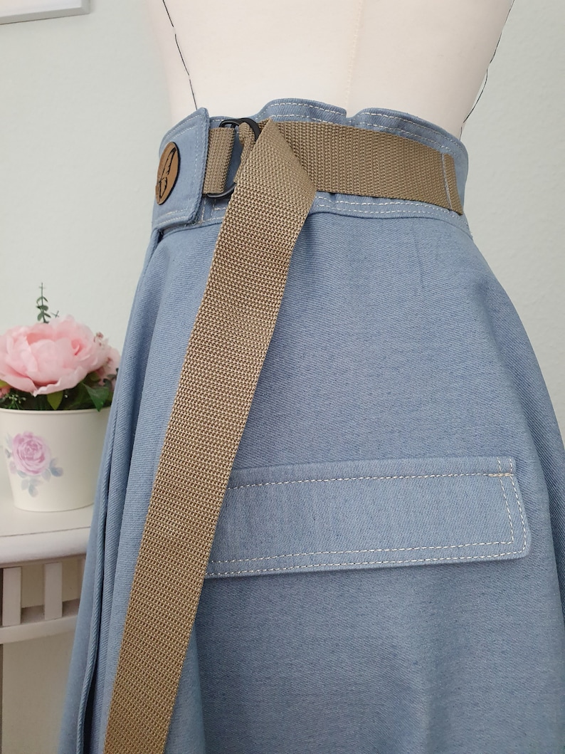 May include: Light blue denim skirt with a tan belt and a pocket detail. The skirt has a high waist design with a decorative wooden button. The tan belt is made of a woven material and has a black buckle.