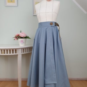 May include: A light blue skirt with a wrap design, featuring a brown belt with a wooden buckle. The skirt is displayed on a mannequin with a floral accent. The skirt has a flowing, flared silhouette.