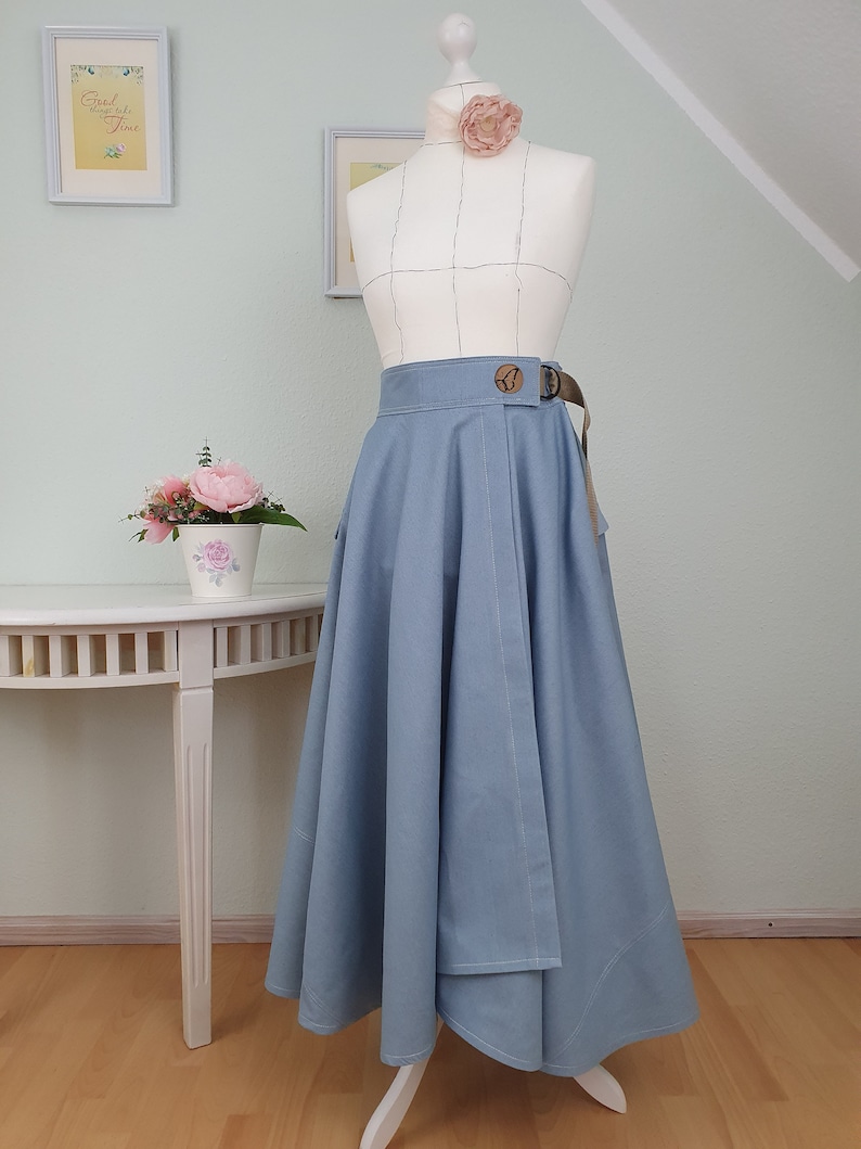 May include: A light blue skirt with a wrap design, featuring a brown belt with a wooden buckle. The skirt is displayed on a mannequin with a floral accent. The skirt has a flowing, flared silhouette.
